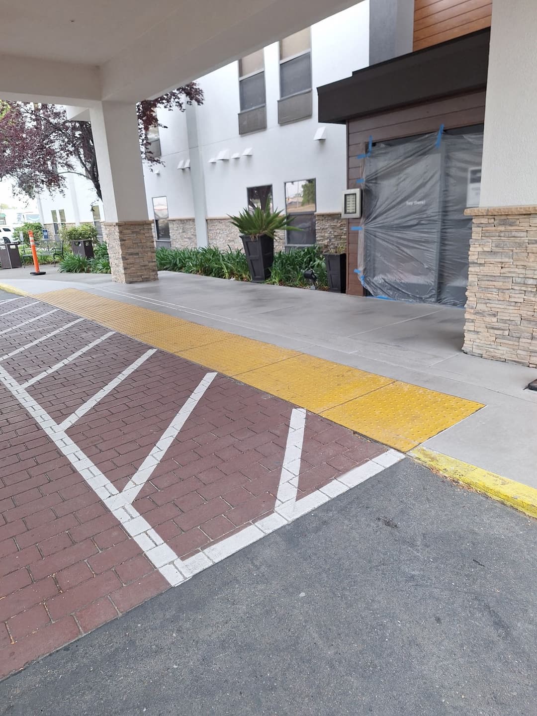 Entrance to a building with a yellow tactile path, brick and concrete paving, and construction barriers.