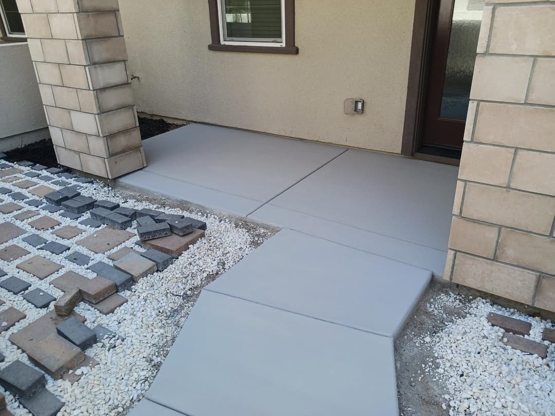 Newly poured concrete walkway leading to a home's entrance, surrounded by paving stones and gravel.