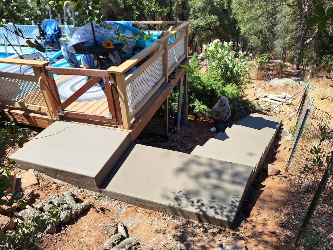 Concrete steps leading to a deck with a pool, surrounded by greenery and trees.