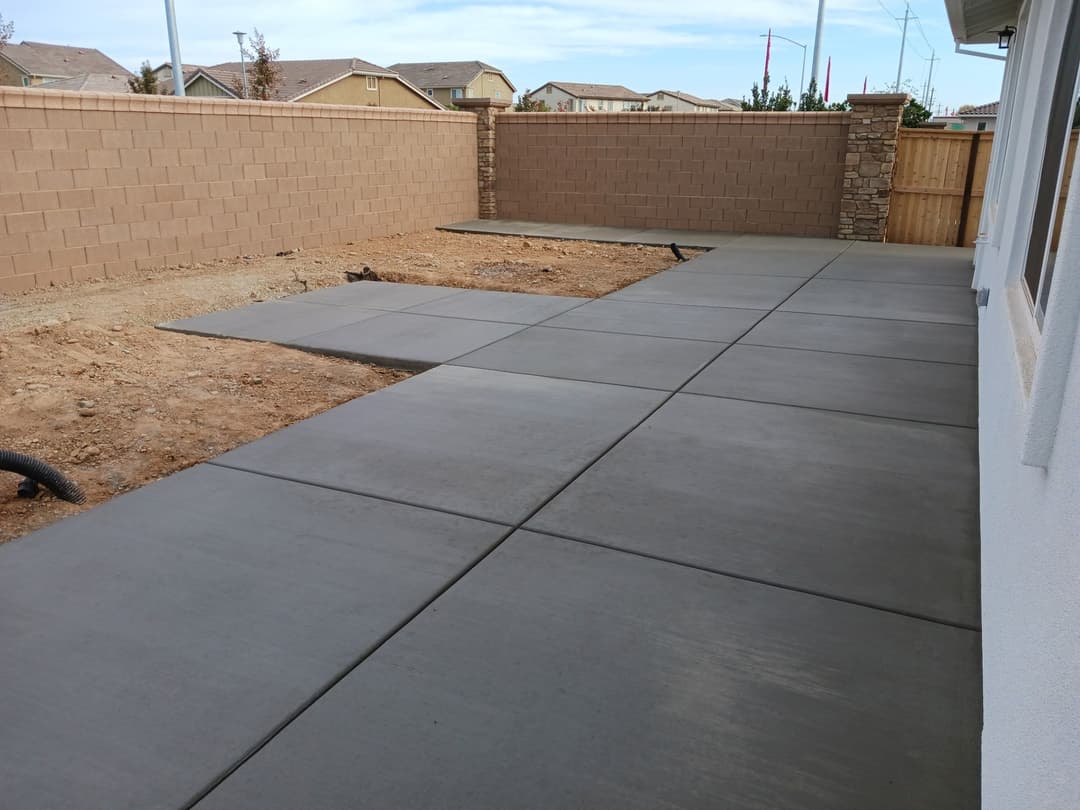 Newly poured concrete patio in a backyard, featuring smooth gray surfaces and a surrounding wall.